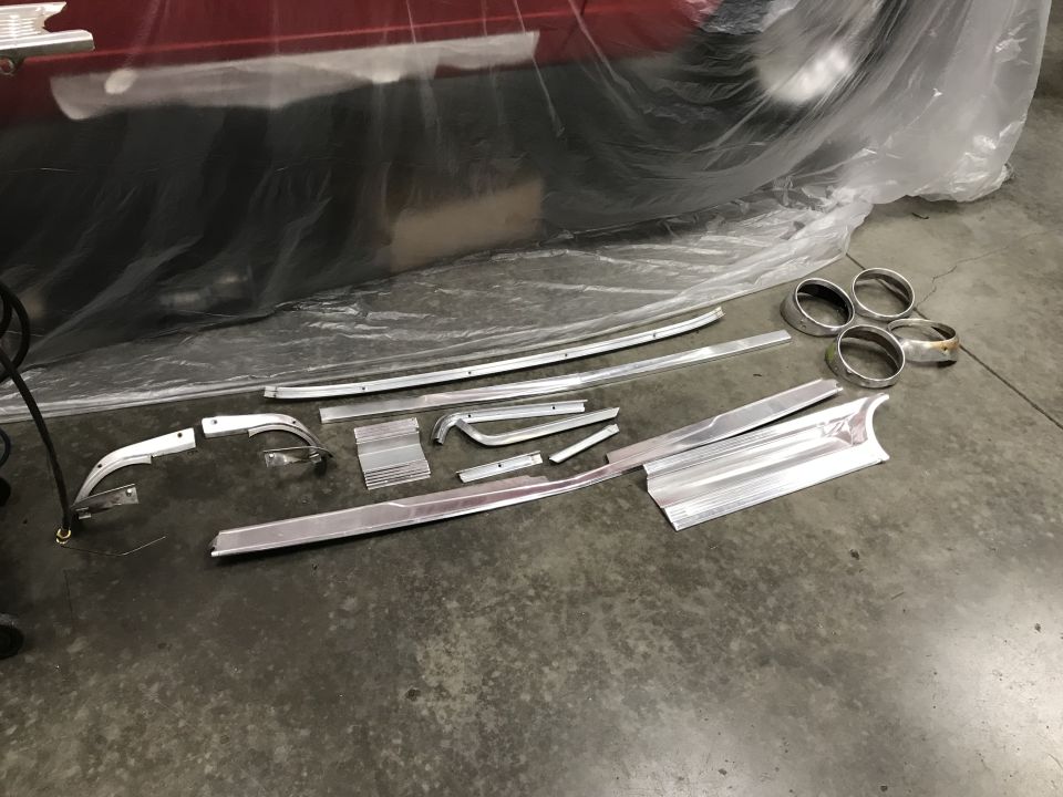 Polished aluminum trim pieces and headlight bezels on concrete floor near a red vehicle covered in plastic.