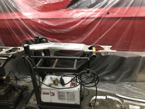 1964 Ford Fairlane - A chrome grille assembly is mounted on a cart in front of a red vehicle covered in plastic sheeting. A welding machine with "ABICOR BINZEL"…