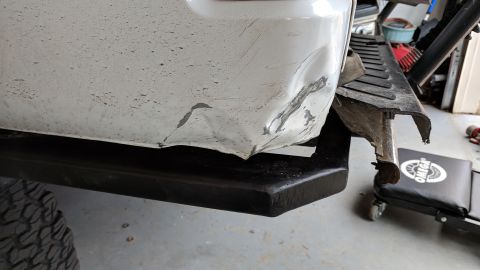 1995 Ford Bronco - A damaged white rear bumper and fender on a vehicle, with a black step bar visible below. The bumper shows significant dents and scrapes, w…