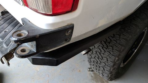 1995 Ford Bronco - A close-up view of the rear corner of a white SUV, showing a black aftermarket bumper with tow hitch receivers and a portion of the treaded…