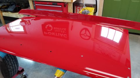 1970 Chevrolet Camaro - A bright red car hood is shown from a high angle, resting on a red stand with a tire visible underneath. The glossy surface reflects a wind…