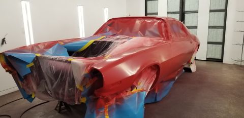 1970 Chevrolet Camaro - A red Chevrolet Camaro is shown in a paint booth, covered in protective plastic and tape. The car is on jack stands and appears to be under…