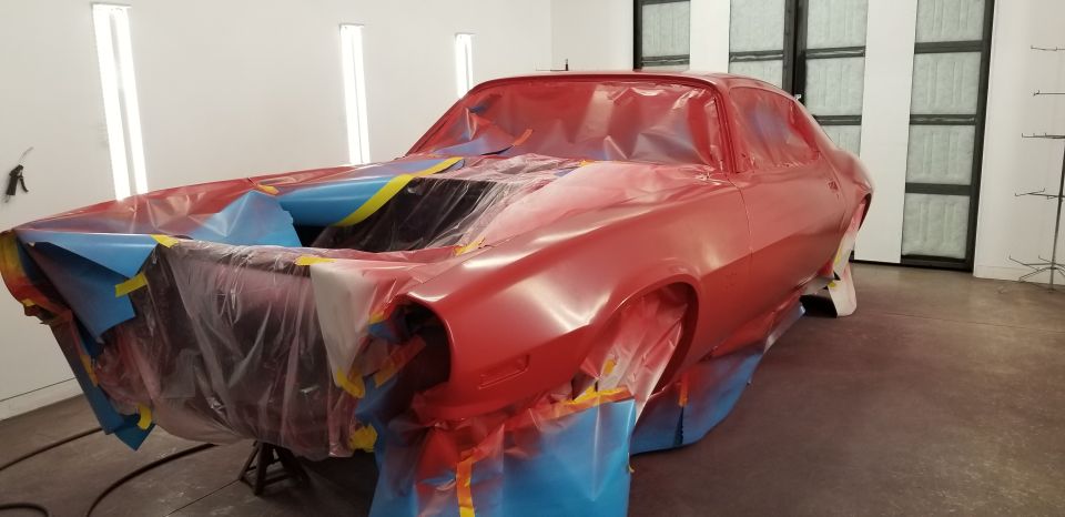 Red 1970 Chevrolet Camaro covered in plastic and tape in a paint booth.