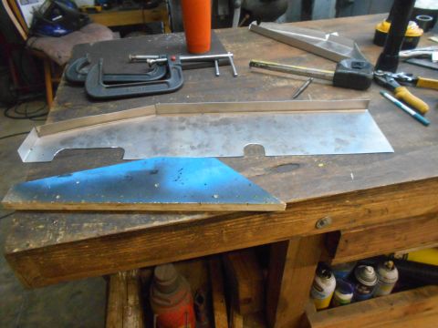 1992 Ford Bronco - A workbench in a workshop is cluttered with tools and metal parts. A long, bent piece of sheet metal and a blue-painted metal piece are lai…