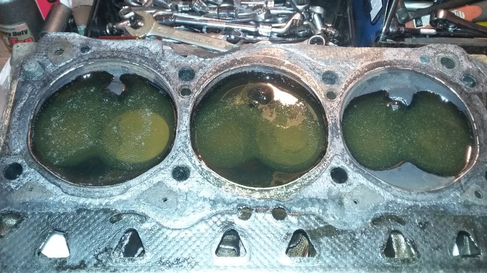 Engine cylinder head with three cylinders filled with green liquid, tools in background