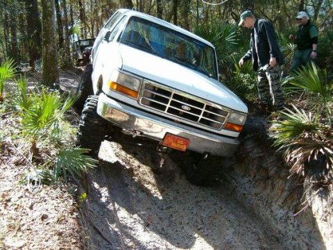 1995 Ford Bronco - White full-size Ford Bronco on a lift with mud-terrain tires tilted up out of a deep sandy off-road rut, two men standing alongside amid pa…