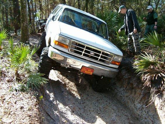 White lifted Ford Bronco climbing a sandy rut off-road with two men beside