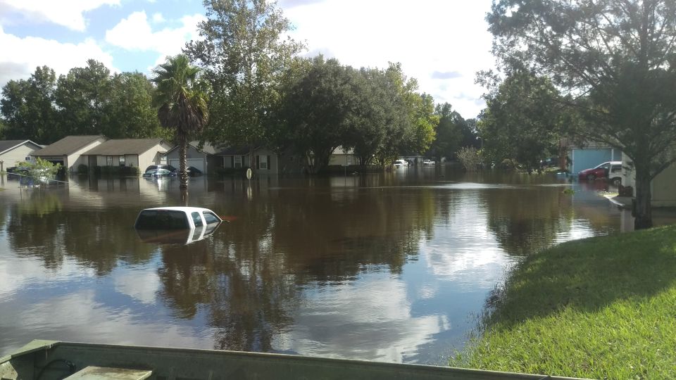 Flooded neighborhood with houses and cars underwater. A white SUV is partially submerged in the foreground