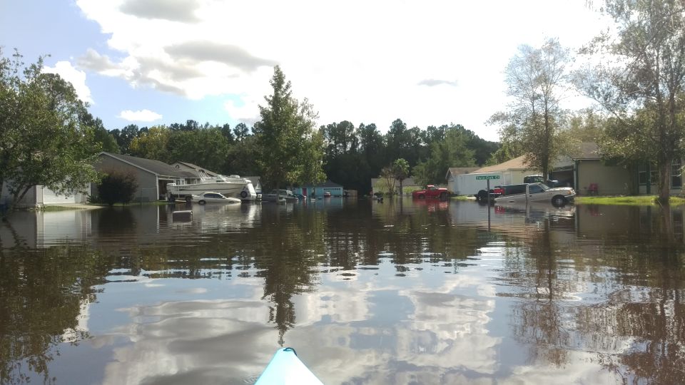 Flooded neighborhood with cars and a boat submerged in water. Kayak in foreground