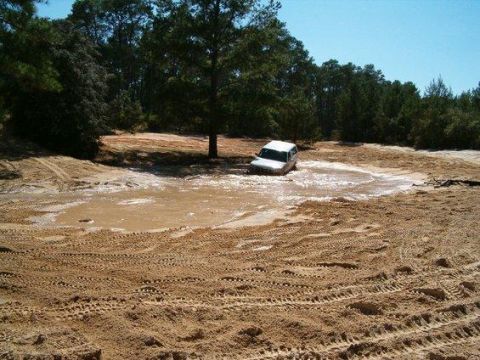 1995 Ford Bronco - A white full-size SUV wades through a wide brown mud pit at a sandy off-road park, water splashing around its wheels and only the upper bod…