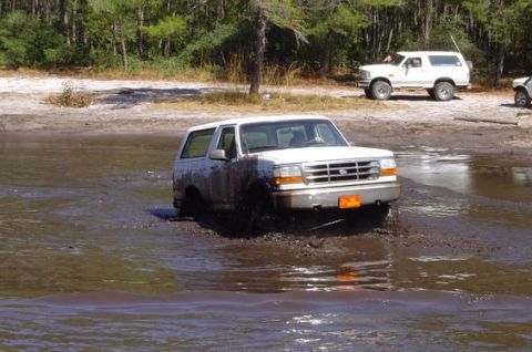 1995 Ford Bronco - A white full-size Ford Bronco driving through a shallow muddy water crossing and throwing spray, seen front three-quarter; a second white B…