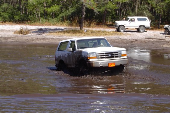 White Ford Bronco fording a muddy water crossing, front 3/4 view.