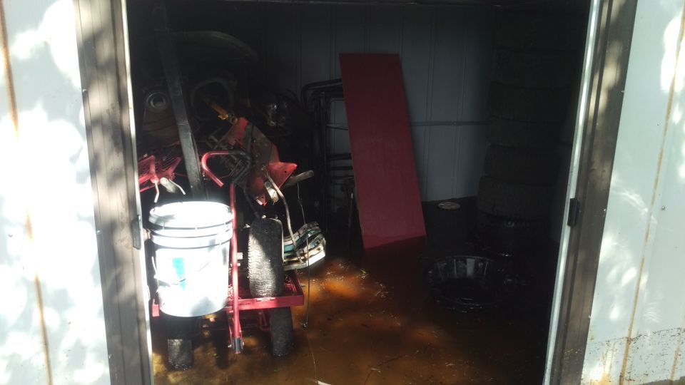 Red garden cart with a white bucket on it, parked inside a flooded shed next to stacked tires