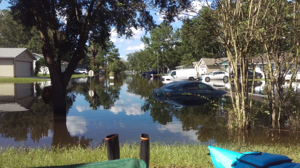 Dark sedan submerged in floodwater on a residential street with houses and trees