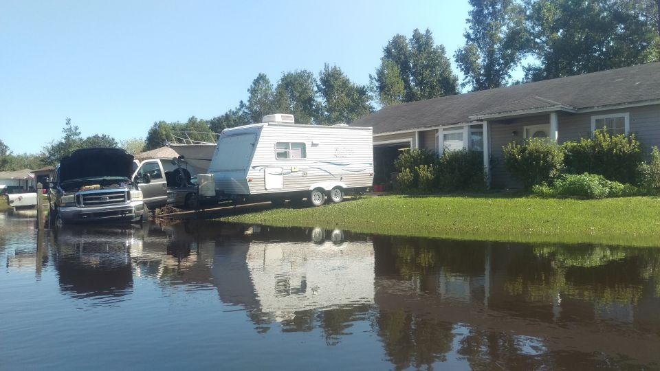 Front view of a dark-colored truck with hood up, partially submerged in floodwaters, with a trailer parked nearby