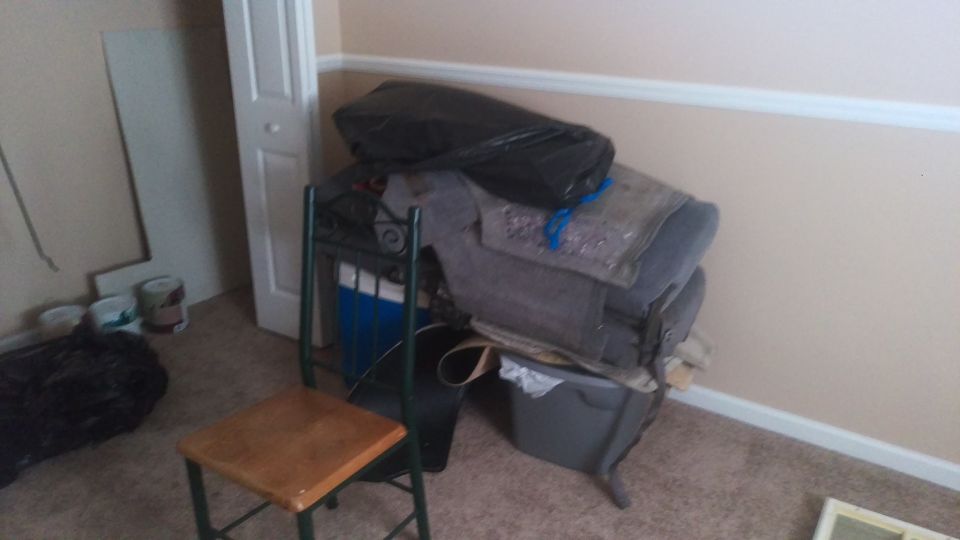 Stacked items including a black trash bag, fabric, and a plastic bin in a room with a chair