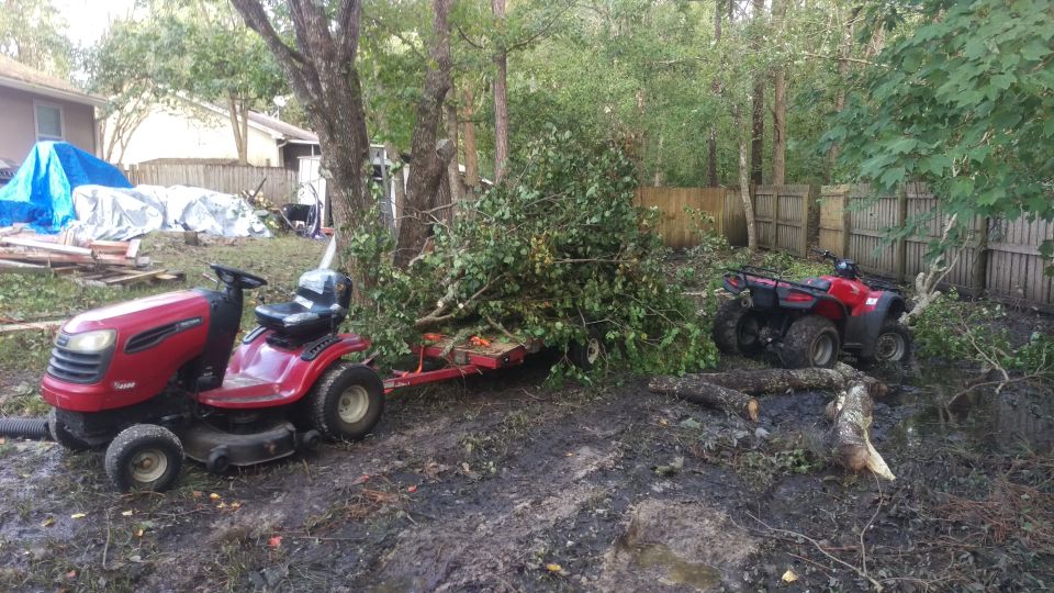 Red riding mower with trailer loaded with branches, and a red ATV in a muddy yard with fallen trees and debris