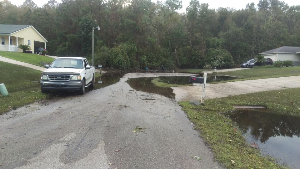 White pickup truck parked on a flooded street in front of a house. Another car is visible in the background