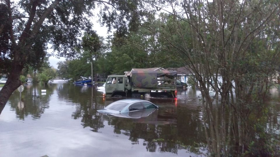 Flooded street with a submerged car and a military-style truck. Houses and trees are visible in the background