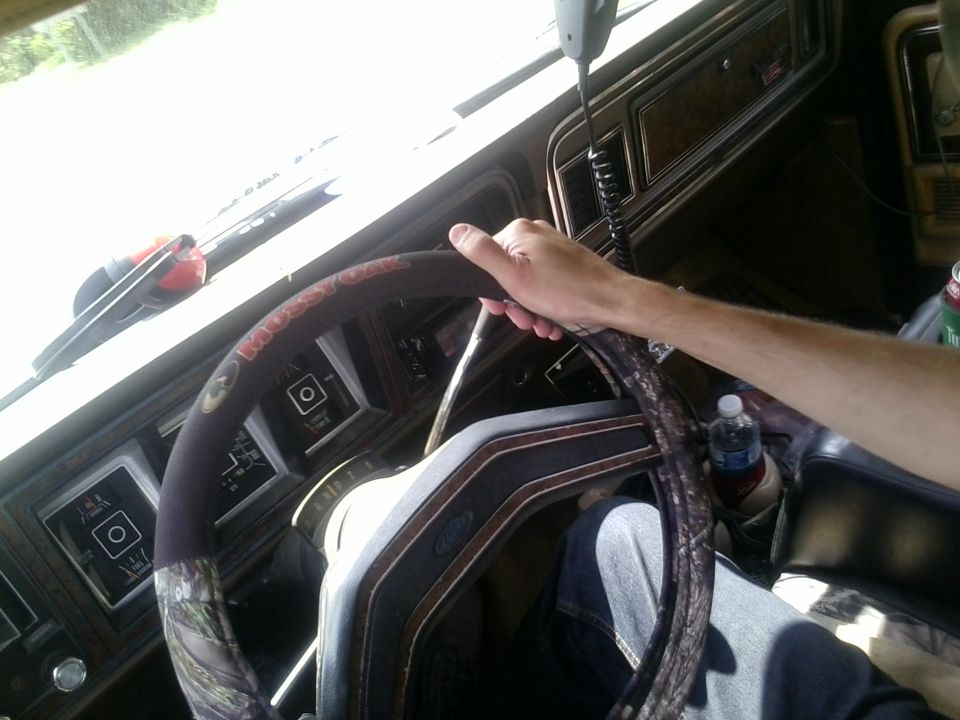 Driver's hand on steering wheel in a 1970s truck interior; wood grain dash and camouflage steering wheel cover visible