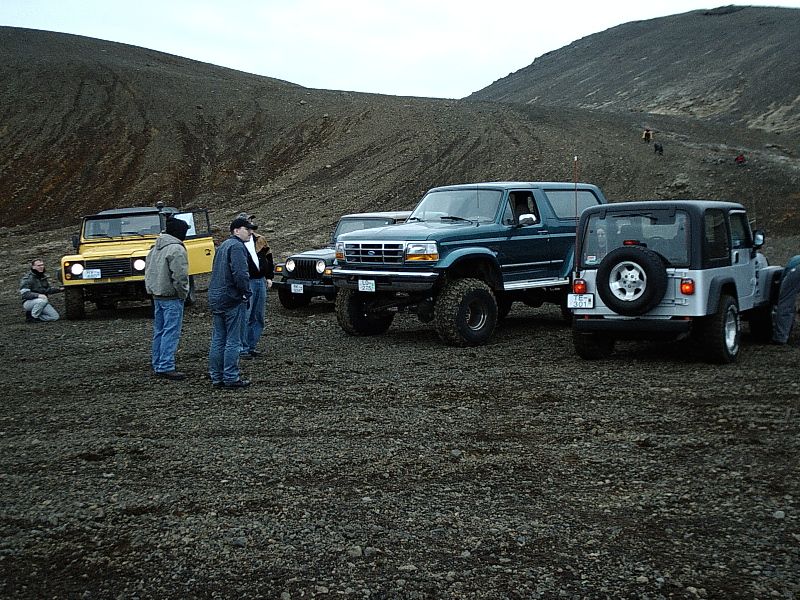 Yellow Bronco, teal lifted Ford, and silver Jeep Wrangler parked on gravel with people