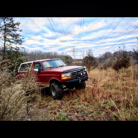 1995 Ford Bronco - This red 1995 Ford Bronco is equipped with a winch and an aftermarket bumper, seen off-road under a cloudy sky.