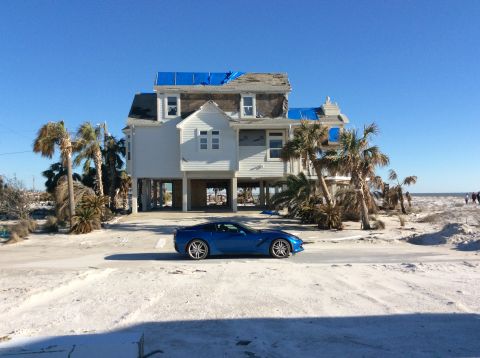 1992 Ford Bronco - A blue sports car is parked on a sandy road in front of a house that appears to have sustained storm damage.