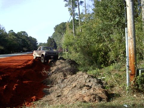 1985 Ford Bronco - A brown pickup truck is positioned on a dirt mound next to a wooded area.