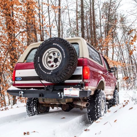 1995 Ford Bronco - A red 1995 Ford Bronco navigates a snowy path surrounded by trees with autumn leaves.