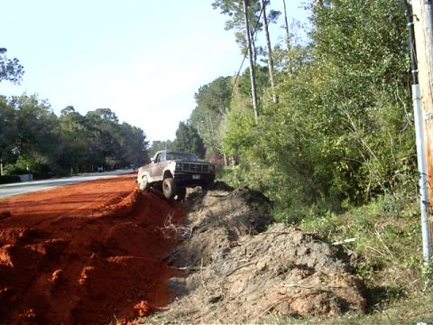 1985 Ford Bronco - A lifted Ford truck navigates a challenging dirt incline beside a paved road.