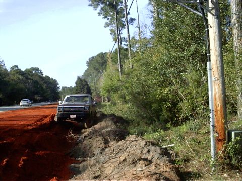 1985 Ford Bronco - A brown Ford F-Series truck is parked off the side of a dirt road next to trees and a utility pole.