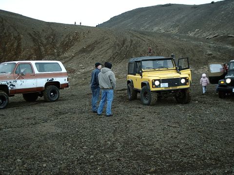 1996 Ford Bronco - A yellow Land Rover Defender 90 is parked on a gravelly terrain with other vehicles nearby.