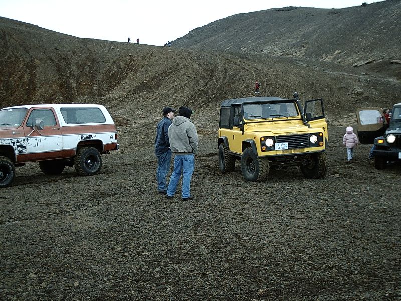 Yellow Defender-style 4x4 and other lifted trucks on gravel with two men talking
