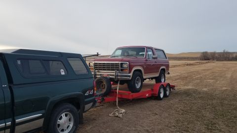 1985 Ford Bronco - A red Ford Bronco is being transported on a trailer, towed by a pickup truck in a rural setting.