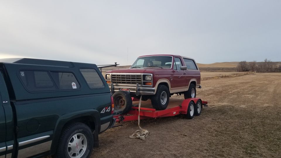 Red Ford Bronco on a red trailer being towed by a dark green pickup truck