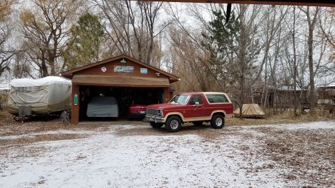 1985 Ford Bronco - A red and tan SUV is parked outside a garage, with a trailer to the left and another vehicle covered inside the garage.