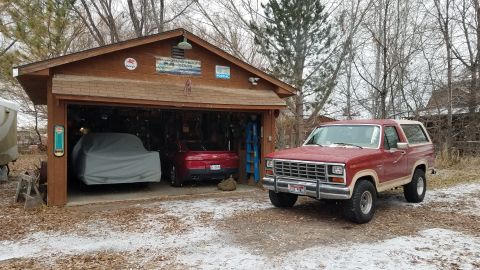 1985 Ford Bronco - A red 1985 Ford Bronco is parked in front of a wooden garage with two cars covered inside.