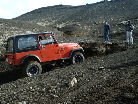 1996 Ford Bronco - An orange Jeep CJ-7 is driven off-road through a rocky, uneven landscape with two people observing.