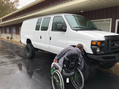 2010 Ford E250 - A white 2010 Ford E250 van with oversized tires is shown from the front 3/4 view, with a person in a wheelchair nearby.