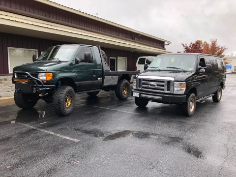 2010 Ford E250 - Two vans are parked in a wet parking lot outside a building.