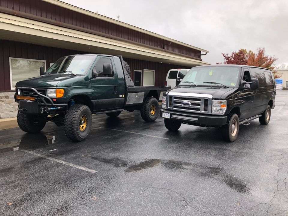 Black Ford E-Series van parked next to a green flatbed truck with large tires
