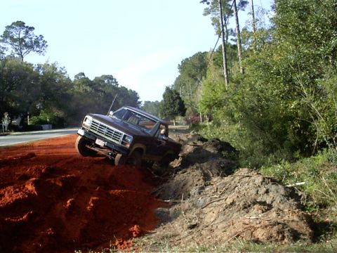 1985 Ford Bronco - A pickup truck navigates a steep, muddy incline next to a wooded area.
