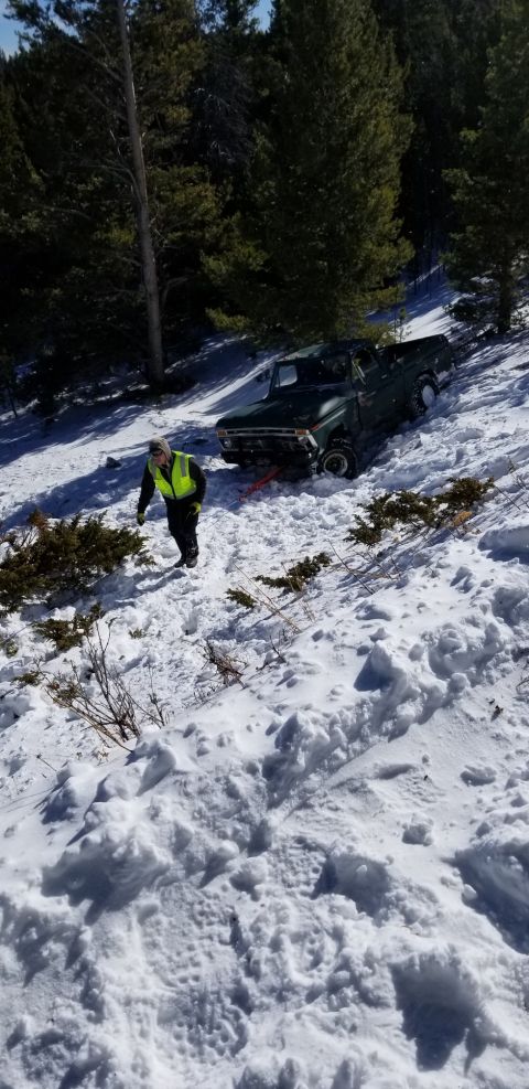 1996 Ford Bronco - A green pickup truck is stuck in the snow on a steep incline with a person in a high-visibility vest standing nearby.