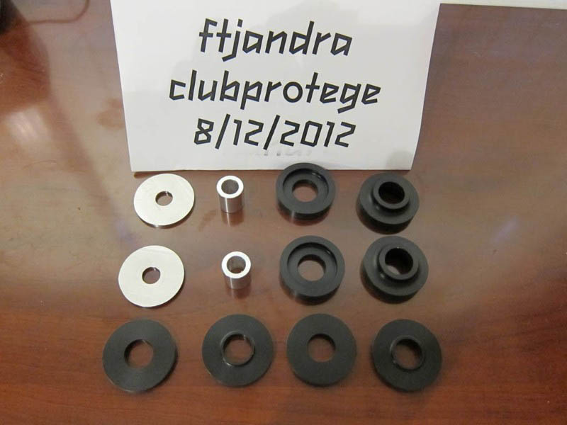 Set of black and silver suspension bushings and metal sleeves on a wooden surface. Paper with text 'ftjandra clubprotege 8/12/2012' behind the parts