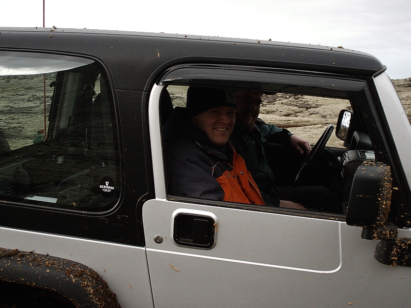 Muddy silver two-door SUV with two smiling men inside, seen from the passenger side