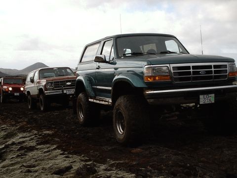 1996 Ford Bronco - A line of SUVs, including a Ford Bronco, navigate a rough terrain under a cloudy sky.