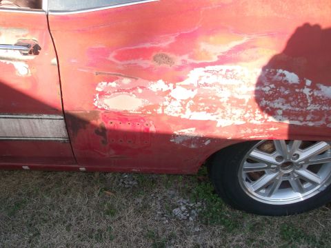 1970 Ford Torino - The side of a car shows extensive body damage and rust, with primer exposed through the red paint.
