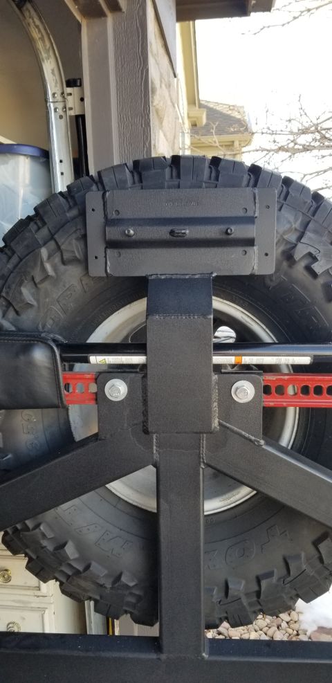 1996 Ford Bronco - A close-up view of a spare tire mounted on a black metal rack, with a Hi-Lift jack secured to the rack. The tire is large and aggressive, s…