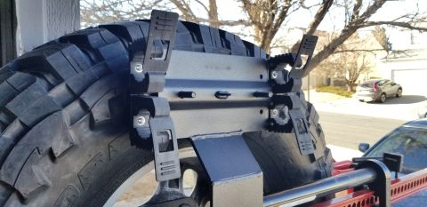 1996 Ford Bronco - A close-up view of a large off-road tire mounted on a black metal spare tire carrier, which is attached to a red Hi-Lift jack. The tire and…