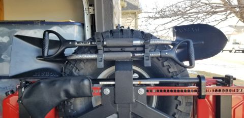 1996 Ford Bronco - A close-up view of two shovels mounted to the spare tire of an off-road vehicle. A red high-lift jack is also visible mounted to the rear o…